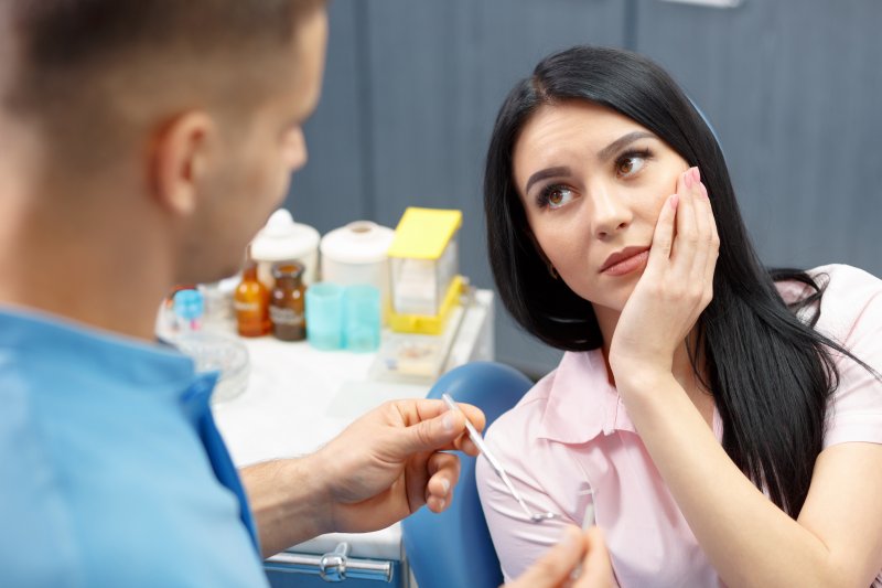 Woman in dentist chair holding cheek while facing dentist holding dental tool